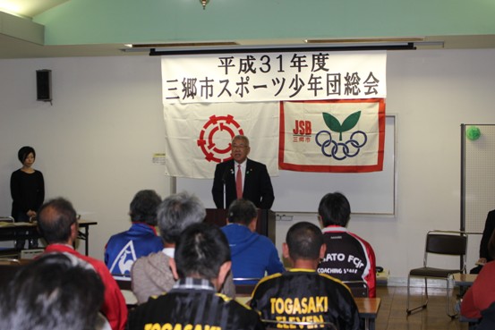 「平成31年度三郷市スポーツ少年団総会」と書かれた紙が設置されている部屋で、様々なデザインのジャージを着用した関係者たちが椅子に着席しているなか、演壇に立つ市長が挨拶をしている写真