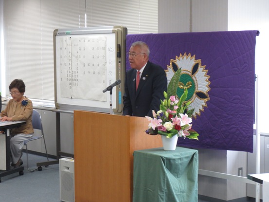 紫色の布地の中心にマークが描かれており、その前に演台や花台が用意された部屋で、演台に立って挨拶をしている市長の写真
