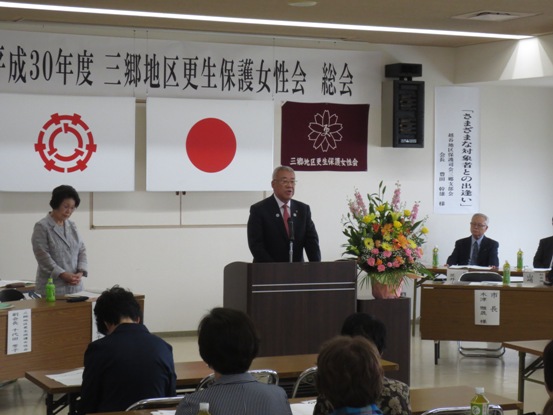 壁に「平成30年度三郷地区更生保護女性会総会」と書かれた紙が設置された部屋で、参加者たちが椅子に座っているなか、正面の演台に立って挨拶をしている市長の写真