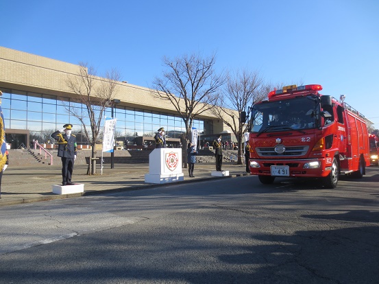 三郷市文化会館の前で三郷市章と消防マークが入った演台に立ち敬礼をしている三郷市長と関係者の方々の前を消防車が通っている写真