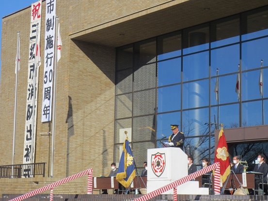 「市制施行50周年 祝 三郷市消防出初式」と書かれた大きな幕が文化会館に設置され、三郷市章と消防マークが入った演台に立ち告辞を述べている三郷市長と長机と椅子が設置された席に座っている関係者の方々の写真