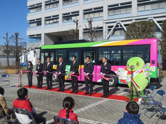 ラッピングバスが置かれた市役所前の会場で三郷市長、6人の関係者の方々、かいちゃんがレッドカーペットの上に立ち、テープカットをしようとしている写真