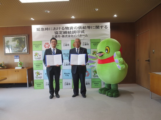 「緊急時における物資の供給に関する協定締結調印式」と書かれた三郷市のバックボードの前で、株式会社ビバホームの男性と三郷市長が協定書を持ち、かいちゃんも一緒に記念撮影をしている写真