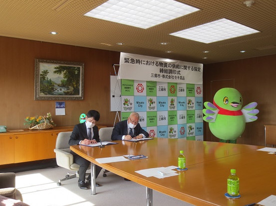 「緊急時における物資の供給に関する協定締結調印式」と書かれた三郷市のバックボードの前で、株式会社セキ薬品の男性と三郷市長がサインをしている写真