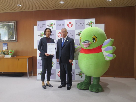三郷市のバックボードが設置された部屋で(左から)PR大使の高橋萌木子さん、三郷市長、かいちゃんが一緒に記念撮影をしている写真