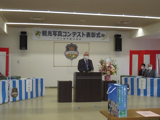 「観光写真コンテスト表彰式」と書かれたプレートと三郷市観光協会会員証マークの前で演台に立ち挨拶をする三郷市長と、白と水色のストライプ柄の敷物が敷かれたテーブルと椅子が設置された席に座って聞いている関係者の方々の写真