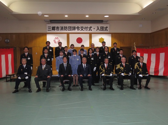「三郷市消防団辞令交付式・入団式」と書かれた横断幕と三郷市章、国旗、消防紋章が掲げられた会場で新入団員、階級昇格者、三郷市長、関係者の方々が一緒に記念撮影をしている写真