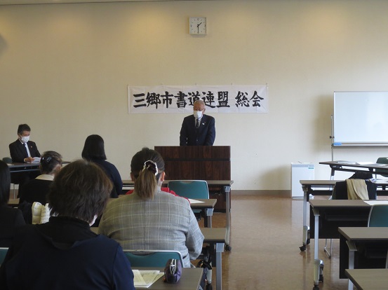 「三郷市書道連盟総会」と書かれた横断幕の前で三郷市長が演台に立って挨拶をし、関係者の方々が席に座って聞いている写真