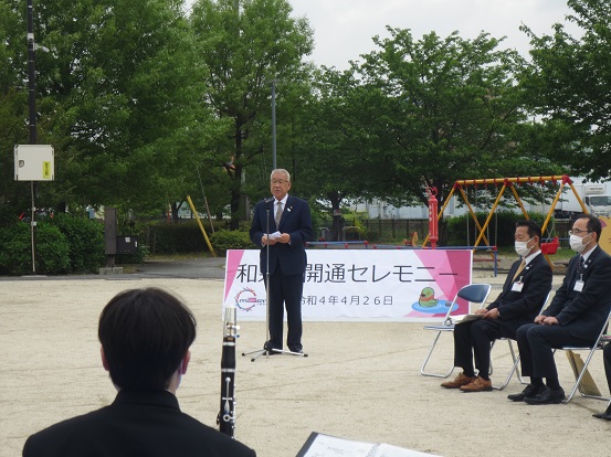 月の郷公園でスタンドマイクの前に立って挨拶する三郷市長と、パイプ椅子に座って聞いている関係者の方々の写真