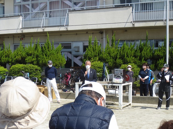 高州東小学校のグラウンドで三郷市長が前方に立って挨拶をし、立って聞いている参加者の方々の後方から写した写真