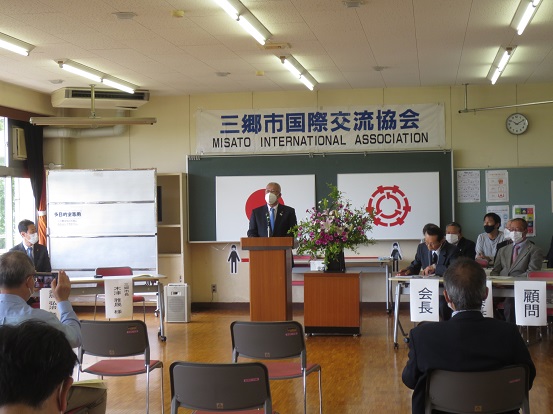 「三郷市国際交流協会」と書かれた横断幕の前で演台に立ち挨拶する三郷市長と、脇に関係者の方々が座っている総会の様子の写真