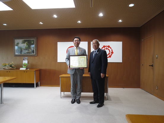 国旗と三郷市章が壁に掛けられた部屋で、株式会社横引シャッター様の男性(左)が賞状を持ち、三郷市長(右)と一緒に記念撮影をしている写真