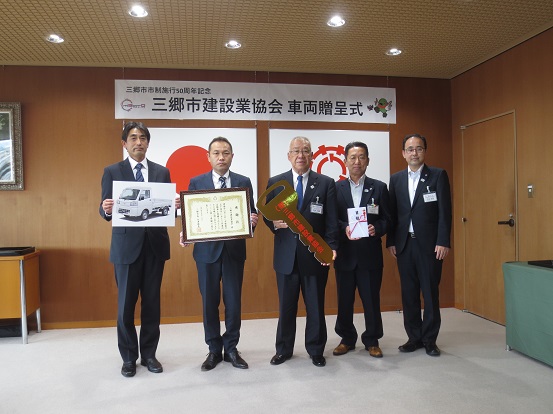 「三郷市建設業協会 車両贈呈式」と書かれた横断幕と国旗、三郷市章が飾られた部屋で、三郷市建設業協会の関係者の方々が賞状や車の写真を持ち、三郷市長がレプリカキーを持って一緒に記念撮影をしている写真