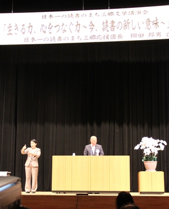 「日本一の読書のまち三郷文学講演会」と書かれた横断幕の下で、スーツ姿の女性が手話をしながら演壇で挨拶を行っている市長の写真