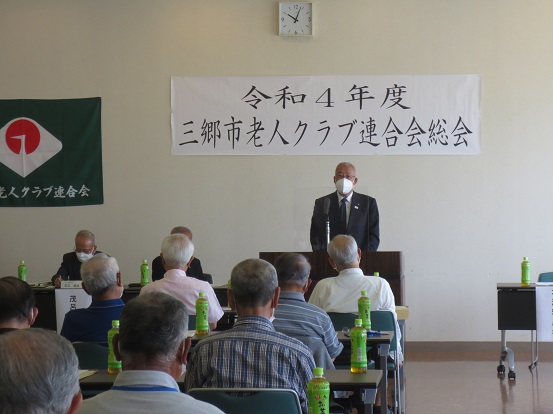 「令和4年度三郷市老人クラブ連合会総会」と書かれた横断幕と老人クラブ連合会の旗が掲げられた会場で演台に立って挨拶する三郷市長と、2名の関係者、たくさんの参加者の方々が席に座って聞いている写真