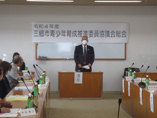 「令和4年度三郷市青少年育成推進委員協議会総会」と書かれた横断幕の前で演台に立って挨拶する三郷市長と、名前プレートが設置された席に座って聞いている関係者の方々の写真