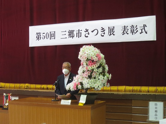 「第50回 三郷市さつき典 表彰式」と書かれた横断幕の前で、白と淡いピンク色のさつきが飾られた演台に立ち挨拶する三郷市長の写真