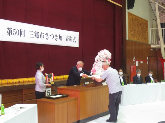 市長賞を受賞した男性が三郷市長から賞状を手渡しされ、サポートの女性がトロフィーを用意している表彰式の様子の写真