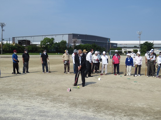 グラウンドでクラブを持ちボールを打とうとしている三郷市長と、見守っているたくさんの参加者の方々の写真