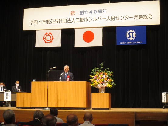 「祝 創立40周年 令和4年度公益財団法人三郷市シルバー人材センター定時総会」と書かれたプレートと三郷市章、国旗、シルバー人材センターの旗が掲げられた会場で、演台に立って挨拶する三郷市長と席に座って聞いている参加者の方々の写真