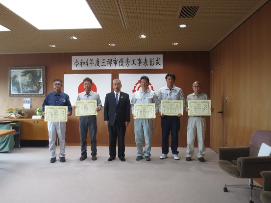 「令和4年度三郷市優秀工事表彰式」の横断幕が設置されている部屋で、市長を中心に表彰をうけた5名の男性が一緒に並んで記念撮影をしている写真