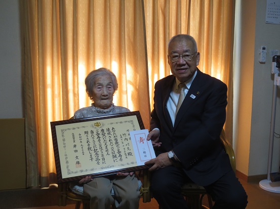 オレンジのカーテンが引かれた部屋で、表彰状を持った竹内いちさんと一緒に、市長が椅子に座って記念撮影をしている写真