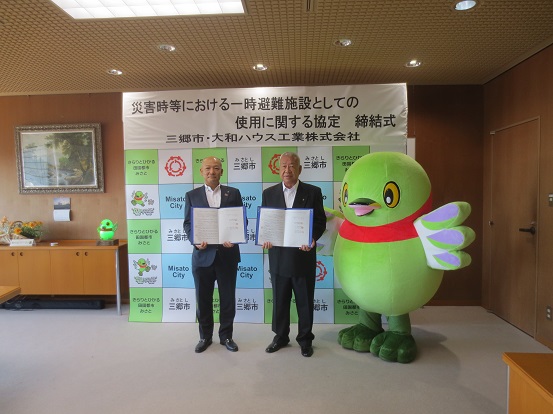 大和ハウス工業株式会社 更科東京本店長と木津市長が協定書を持っており、かいちゃんと一緒に三郷市のバックボードの前に立って記念撮影をしている写真