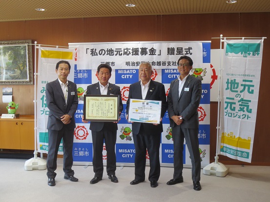 「「私の地元応援募金」贈呈式」の横断幕が設置された三郷市のバックボードの前で、表彰状を持った関係者と市長を中心に、両側に2名の関係者たちが並んで記念撮影をしている写真