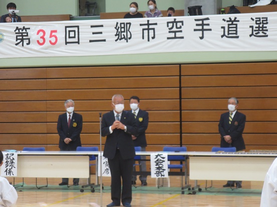 「第35回三郷市空手道選手権大会」の横断幕が下げられた体育館で、マイクを手に挨拶をしている木津市長の写真