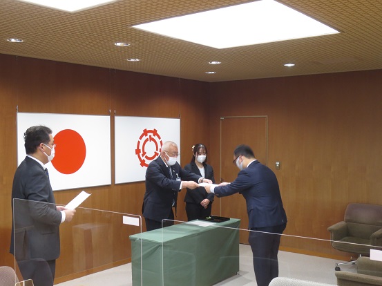 国旗と市章が飾られた室内にて、緑色のテーブルの上で選任書を両手で手渡しする木津市長と受け取る公平委員の方、見守る関係者2名の写真