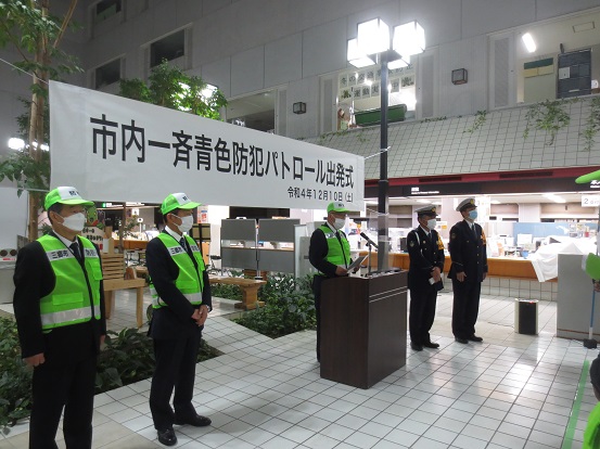 日が落ちた屋外で、市内一斉青色防犯パトロール出発式の横断幕が掲げられた前で、黄緑色の帽子や反射ベストを着用し、講演台で挨拶をする木津市長と警察官2名、自主防犯団体2名の写真