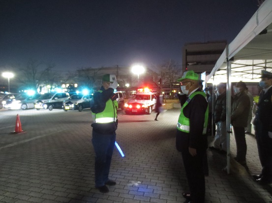赤い回転灯のパトカーと、青の回転灯の自主防犯パトロールカーが整列する前で、敬礼をしながら挨拶を取り交わす反射材ベストを着用した関係者と木津市長の写真