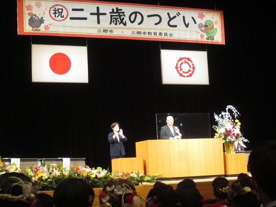 両端にかいちゃん、つぶちゃんが描かれた祝二十歳のつどい横断幕や国旗、市章の下、大きな生花が飾られた壇上で挨拶をする木津市長と手話通訳者の方の写真