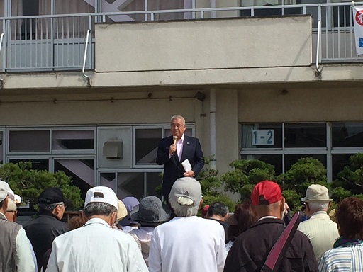 校舎の前に設置された朝礼台の上に乗り右手にマイクを持ち挨拶を行っている市長の話を、並んで聞いている参加者の後ろ姿の写真