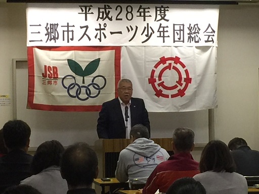 「平成28年度三郷市スポーツ少年団総会」と書かれた横断幕と2枚の旗が飾られた室内に設置された長机の席に座っている参加者と、向かい合って演壇に立っている市長が挨拶を行っている写真