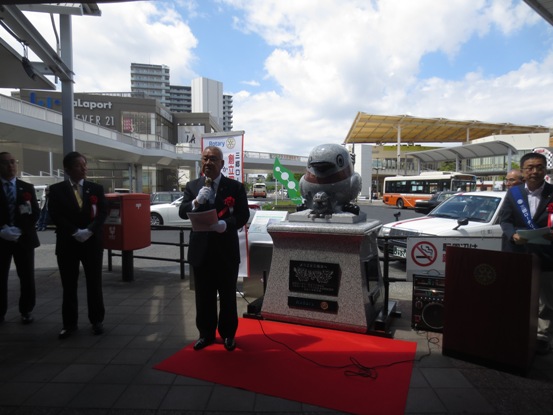 かいちゃんとつぶちゃんの石像が駅前に設置され、石像前に敷かれたレッドカーペット上に立ってマイクを手に話をしている市長の写真