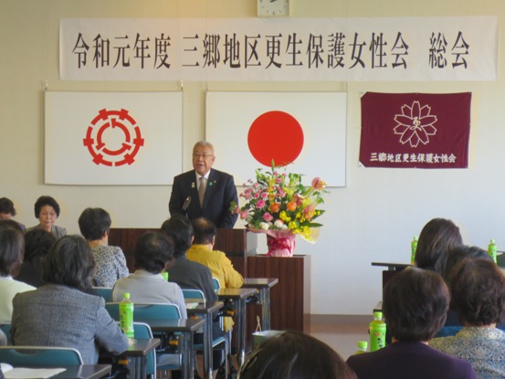 「令和元年度三郷地区更生保護女性会総会」と書かれた紙が貼られている部屋で、椅子に着席している多くの女性たちと、演壇に立って話をしている市長の写真