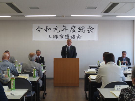 「令和元年度総会三郷市遺族会」と書かれた紙が壁に貼られている部屋で、関係者たちが椅子に着席しているなか、演壇に立つ市長が挨拶をしている写真