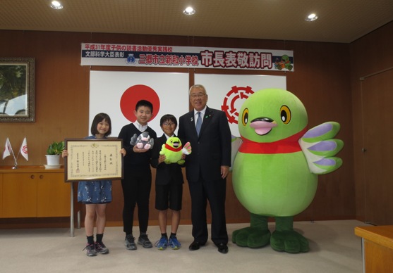 「三郷市立新和小学校市長表敬訪問」と書かれた紙が貼られた部屋で、左から表彰状を持った女の子、つぶちゃんマスコットを持った男の子、かいちゃんマスコットを持った男の子、市長、かいちゃんが並んで記念撮影をしている写真