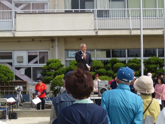 校庭にたくさんの参加者たちが集まっているなか、設置された舞台に立ってマイクを手に話をしている市長の写真