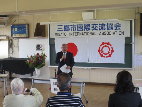 「三郷市国際交流協会」の紙が黒板に貼られている部屋で、関係者たちが正面に立って話をしている市長の方を見ている写真