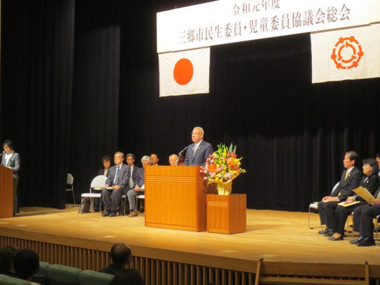 「令和元年度三郷市民生委員・児童委員協議会」と書かれた吊り看板が設置されている舞台上で、関係者たちが両側に着席しているなか、演壇に立って話をしている市長の写真
