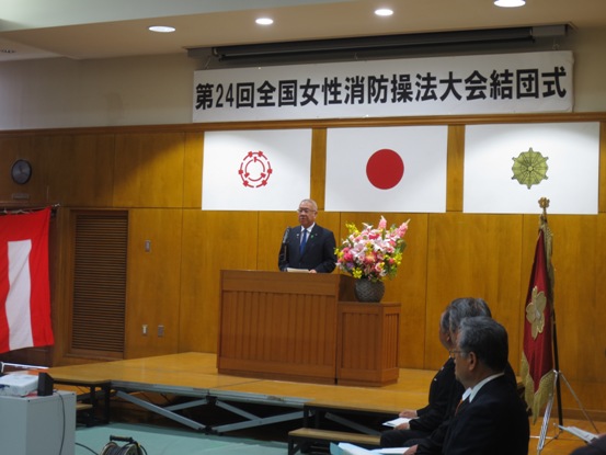 「第24回全国女性消防操法大会結団式」と書かれた吊り看板が設置された部屋で、演壇に立って話をしている市長と、舞台下で市長の方を向いている関係者たちの写真