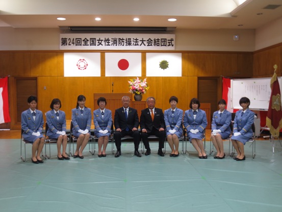 「第24回全国女性消防操法大会結団式」の吊り看板が設置された部屋で、市長と関係者を中心に、両側に4名ずつの女性が水色の制服を着て座っている写真