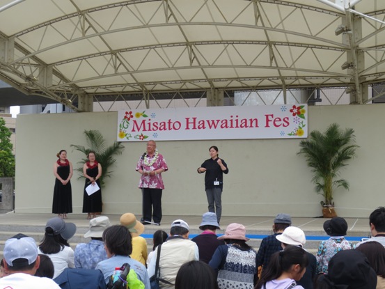 花のイラストで装飾された三郷ハワイアンフェスの看板が屋外の会場に設置され、来場者たちが舞台下に集まっており、ピンク色のシャツとレイを身に着けた市長が舞台上に立って話をしている写真