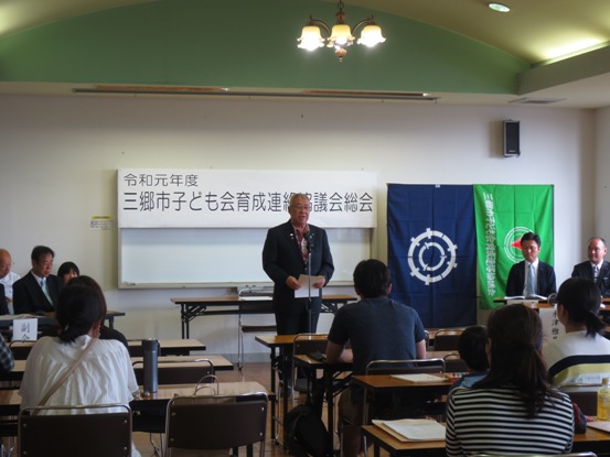「令和元年度三郷市子ども会育成連絡協議会定期総会」のボードが設置された部屋で、椅子に着席している関係者たちと、正面のマイク台の前に立って話をしている市長の写真