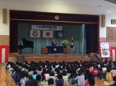 「祝創立40周年記念式典並びご祝賀会」と書かれた横断幕と三郷市旗や国旗が飾られたステージの檀上に立つ市長の話を体育座りをして聞いている児童達の後ろ姿の写真