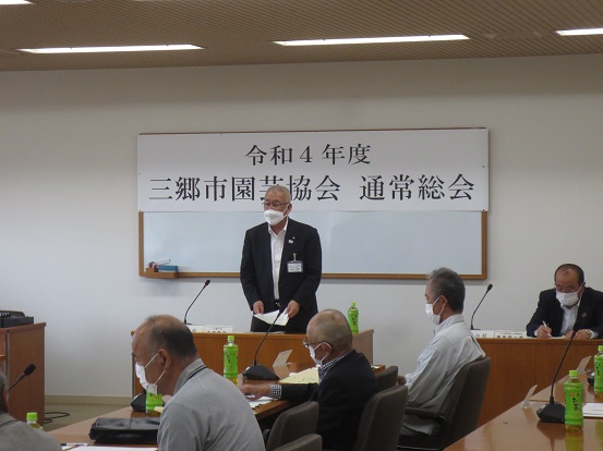 「令和4年度三郷市園芸協会通常総会」と書かれた横断幕の前で席を立って書類を持って挨拶する三郷市長と、席に座って聞いている関係者の方々の写真