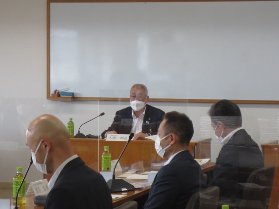 パーテンションで区切られた会議室で三郷市長が座って挨拶をし、名前プレートが設置された席に座って聞いている参加者の男性の写真