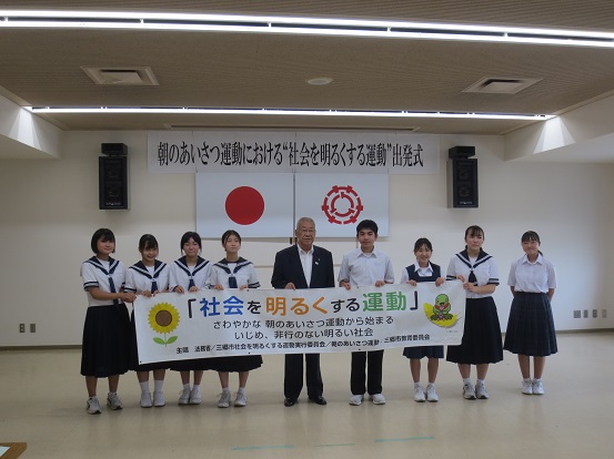 「朝のあいさつ運動における 社会を明るくする運動 出発式」の横断幕が上部に設置されている部屋で、「社会を明るくする運動」の方針が書かれている横断幕を8名の生徒と一緒に持っている市長の写真